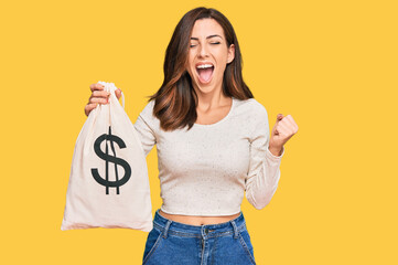 Young brunette woman holding dollars bag screaming proud, celebrating victory and success very...