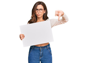 Young brunette woman holding blank empty banner with angry face, negative sign showing dislike with...