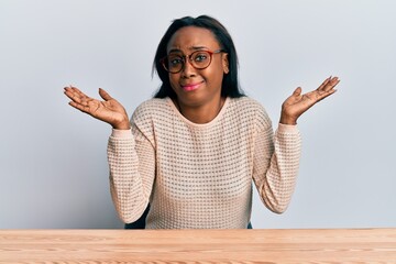 Young african woman wearing casual clothes sitting on the table clueless and confused with open...