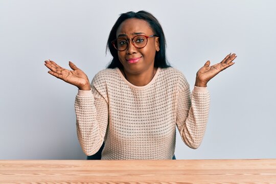 Young african woman wearing casual clothes sitting on the table clueless and confused with open arms, no idea and doubtful face.