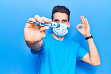 Young handsome man with beard wearing medical mask holding thermometer doing ok sign with fingers, smiling friendly gesturing excellent symbol