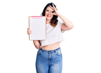 Beautiful young brunette woman holding clipboard with blank space smiling happy doing ok sign with hand on eye looking through fingers