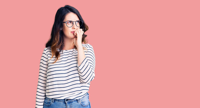 Beautiful young brunette woman wearing casual clothes and glasses looking stressed and nervous with hands on mouth biting nails. anxiety problem.