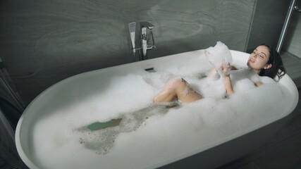 Cute girl lying in bath indoors. Sweet lady enjoying bubble bathtub.