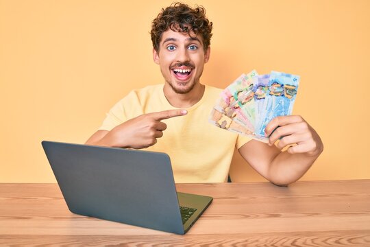 Young caucasian man with curly hair sitting on the table working with laptop and holding canadian dollars banknotes smiling happy pointing with hand and finger