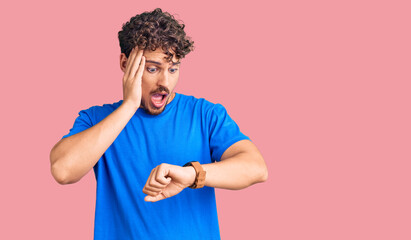 Young handsome man with curly hair wearing casual clothes looking at the watch time worried, afraid...
