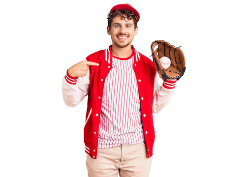 Young handsome man with curly hair wearing baseball uniform holding golve and ball pointing finger to one self smiling happy and proud