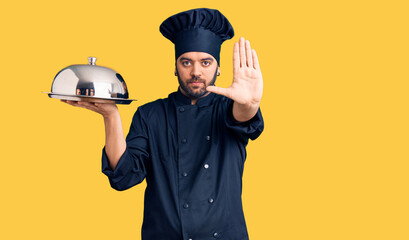 Young hispanic man wearing cooker uniform holding tray with open hand doing stop sign with serious and confident expression, defense gesture