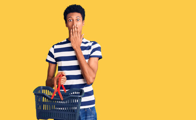 Young african american man holding supermarket shopping basket covering mouth with hand, shocked and afraid for mistake. surprised expression