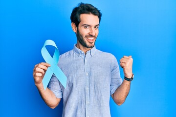 Young hispanic man holding blue ribbon pointing thumb up to the side smiling happy with open mouth