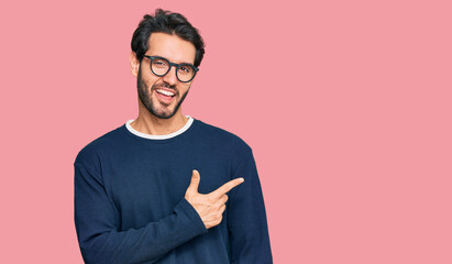 Young hispanic man wearing casual clothes and glasses cheerful with a smile of face pointing with hand and finger up to the side with happy and natural expression on face