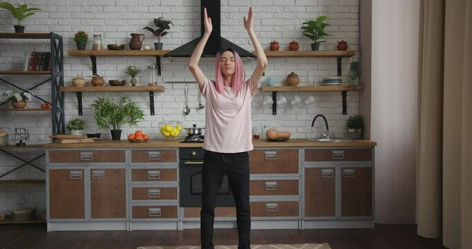 Tranquil transwoman in pink wig and t-shirt does yoga position standing in decorated kitchen with new furniture and equipment