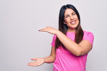 Young beautiful brunette woman wearing casual pink t-shirt standing over white background gesturing with hands showing big and large size sign, measure symbol. Smiling looking at the camera. Measuring