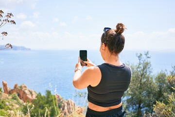 Young hispanic woman on back view wearing sportswear. Making photo using smartphone standing over beautiful landscape