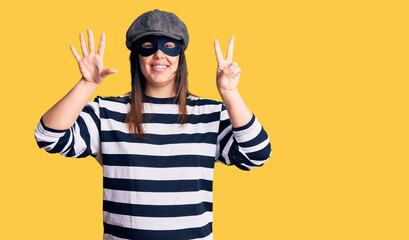 Young beautiful brunette woman wearing burglar mask showing and pointing up with fingers number seven while smiling confident and happy.