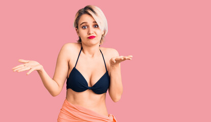 Young beautiful blonde woman wearing bikini clueless and confused expression with arms and hands raised. doubt concept.