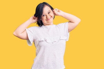 Brunette woman with down syndrome wearing casual white tshirt smiling pulling ears with fingers,...
