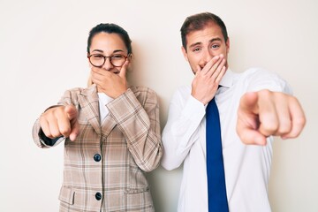 Beautiful couple wearing business clothes laughing at you, pointing finger to the camera with hand over mouth, shame expression