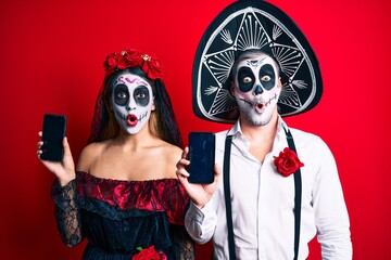 Couple wearing day of the dead costume holding smartphone showing screen scared and amazed with open mouth for surprise, disbelief face