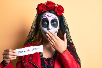 Woman wearing day of the dead costume holding trick or treat paper covering mouth with hand, shocked and afraid for mistake. surprised expression