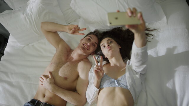 Sexy couple lying on hotel bed. Gorgeous man woman taking selfie mobile phone.