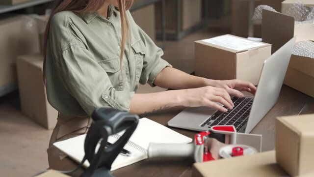 Female entrepreneur online store owner using laptop at work preparing parcel boxes checking ecommerce post shipping online retail e commerce store order fulfillment in dropshipping delivery warehouse.