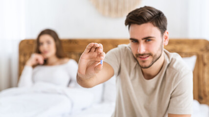 Focus on blue pill in hands of happy young man, and smiling wife sits in bed in modern interior of bedroom