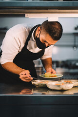Portrait of a male chef with mask decorating food in ceramic dishes over stainless steel worktop in restaurant kitchen during coronavirus pandemic.