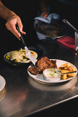 Close-up of the hands of a male chef serving steak with sauce and potatoes in ceramic dish over stainless steel worktop in restaurant kitchen.