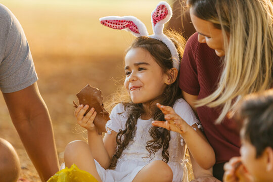 Latin Family Celebrating Easter. Curly Girl Wearing Bunny Ears And Eating Delicious Chocolate Egg