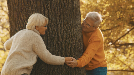 Old couple having romantic autumn day in forest. Hugging tree and smiling. High quality photo