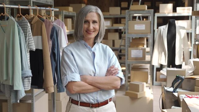 Confident happy mature older 60s woman retail seller, entrepreneur, clothing store small business owner, supervisor looking at camera standing arms crossed in delivery shipping warehouse, portrait.