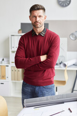 Fototapeta premium Vertical portrait of mature bearded architect standing with arms crossed and looking at camera while posing by drawing desk at workplace