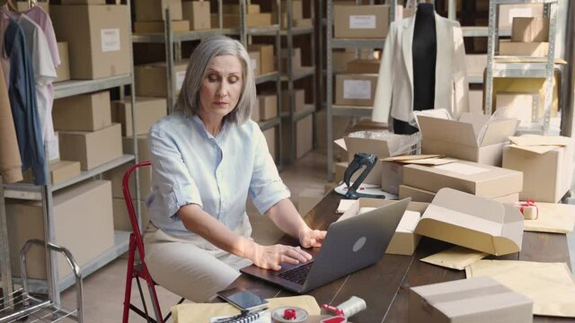 Older female fashion dropshipping business owner using laptop talking on phone in warehouse. Mature woman seller merchant checking online website order confirming ecommerce shipping parcel delivery.