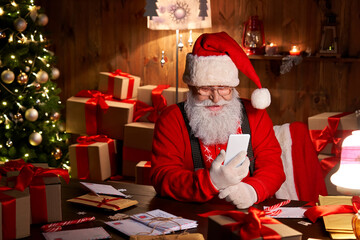 Happy Santa Claus holding smartphone holding cell phone using mobile app for social distance communication, watching videos or video calling in virtual online chat sit at home table late on xmas eve.