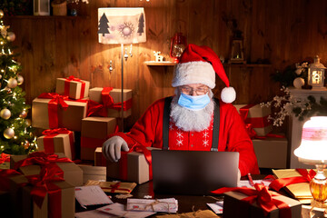 Old bearded Santa Claus wearing face mask, holding gift box on xmas eve sitting at cozy home table...