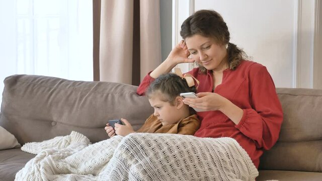 Smiling young mommy and little daughter use contemporary mobile phones sitting under knitted blanket on sofa in living room
