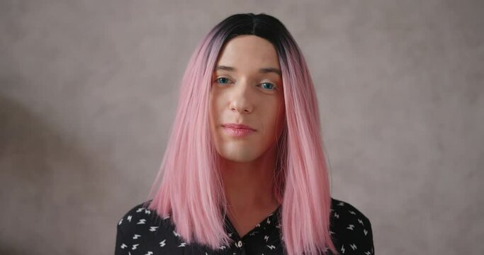 Positive young transwoman wearing black dot blouse and bright pink wig talks posing for camera on beige color background closeup