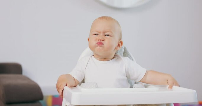 Portrait of young baby in white high baby chair who makes funny faces and grimaces. White walls on background.