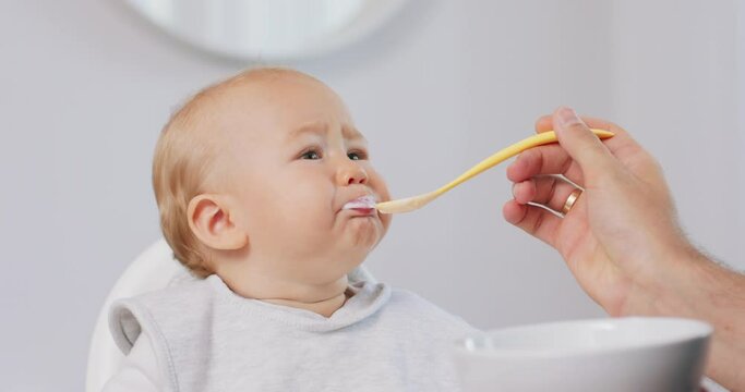 Closeup of young baby in white high baby chair and father's hand with a spoon, while he is feeding the baby, who eats and makes funny grimaces. White walls on background.