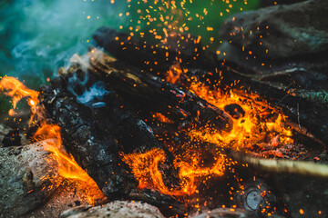 Vivid smoldered firewoods burned in fire close-up. Atmospheric background with orange flame of campfire and blue smoke. Warm full frame image of bonfire. Glowing embers in air. Bright sparks in bokeh