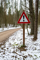 Road sign triangular shape with exclamation mark. Red with white attention traffic sign. Winter road in forest on background.
