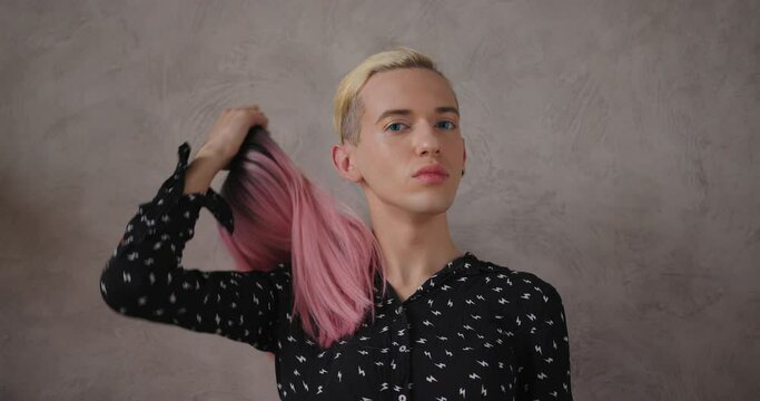 Elegant young transwoman wearing stylish black dot dress takes off bright pink wig posing for camera on beige background closeup