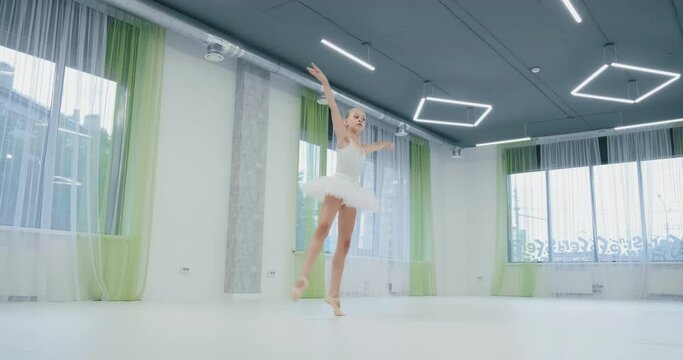 little ballerina in white suit with tutu performs classical dance in studio with large windows