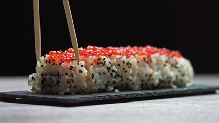 Sticks Take Sushi from Sushi box. Variety of types sushi with red caviar, fish, Philadelphia cheese and chopsticks close-up. Set of delicious Japanese sushi rolls.sushi on a stone board on a black