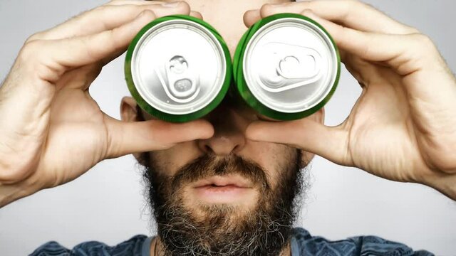 A young bearded man holds two cans of beer to his eyes pretending to look through binoculars