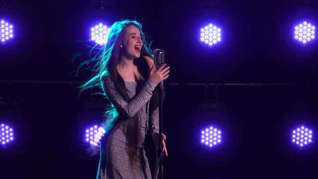 Portrait of a beautiful singer with a vintage microphone on a background of blue lights. Young woman singing a song into a microphone on an empty stage close up. Long hair flying in the wind.