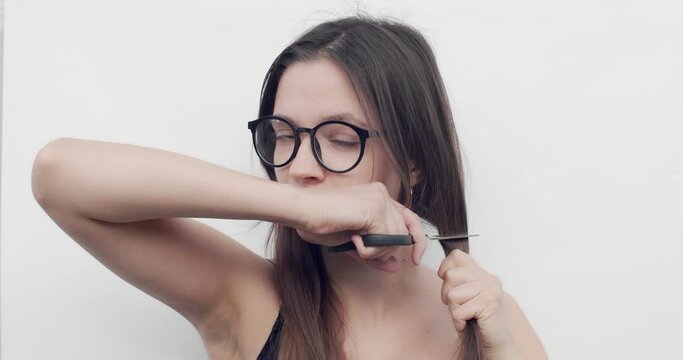 Woman With Glasses Cuts Her Hair On White Background. Young Female Cuts Her Long Brown Hair With Scissors By Herself During Self-isolation. Change Hairstyle For Stress Overcome And Hair Care Concept