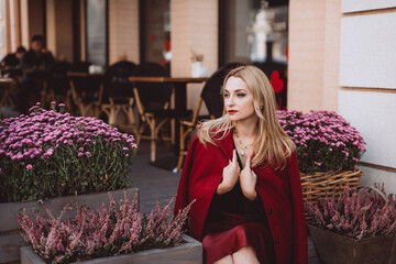 A beautiful young blonde woman in a trendy autumn outfit in burgundy Marsala on the steps of a cafe with chrysanthemums. Soft selective focus. Artistic noise.