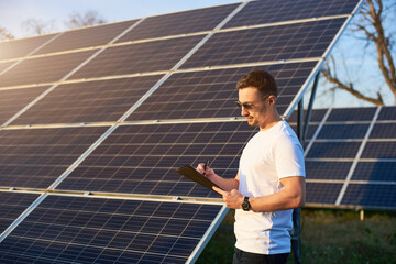 Portrait of a young successful man standing by a solar pv plant looking in his folder pondering on...